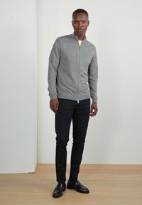 Grå zip-up tröja med hög krage, bärs över en vit skjorta. Svarta slim-fit byxor och glänsande svarta skor kompletterar outfiten.