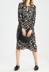 Robe noire à motifs floraux avec des fleurs blanches et jaunes, dotée de manches longues et d'un décolleté en forme de trou de serrure, accompagnée d'une pochette texturée noire.