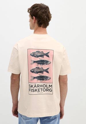 Homme portant un t-shirt beige avec un graphisme de quatre poissons noirs sur fond rose et le texte "Skärholm Fisketorg" dans le dos.