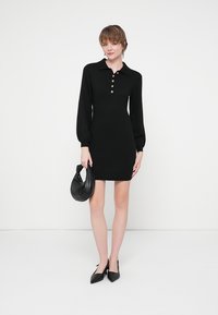 Svart stickad klänning med krage, som har en knappslå och lång ärm. Bärs med en svart kuverttväska och spetsiga klackar.