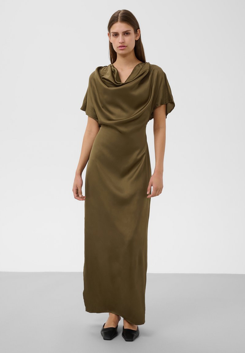 Femme portant une longue robe en satin vert olive avec un décolleté drapé et des manches courtes, debout sur un sol gris devant un fond blanc.
