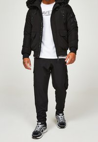 Czarna pikowana kurtka z futrzanym kapturem, zamkiem błyskawicznym, noszona na białym T-shircie, w zestawieniu z czarnymi spodniami cargo i czarnymi sneakersami.
