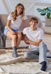 Dos personas están sentadas sobre una manta texturizada de color claro. Una lleva una camiseta blanca y pantalones cortos azules, y la otra, una camiseta blanca y pantalones de pijama a rayas. Ambos llevan calcetines de color gris oscuro.