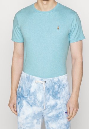 Mężczyzna w jasno-niebieskim t-shircie z krótkim rękawem i białych oraz niebieskich szortach tie-dye z wiązanym paskiem w talii.