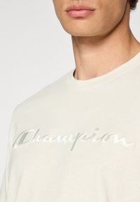 Cremfarbener T-Shirt aus weichem Baumwollstoff mit einem großen, glänzenden, gestickten "Champion"-Logo in hellgrün auf der Brust.