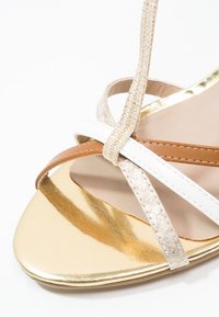 Gros plan d'une sandale dorée avec des brides croisées beige, blanche et marron sur fond blanc.