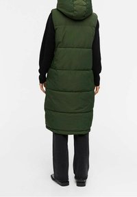 Lange, grüne Steppweste mit Kapuze. Mit gesteppten Abschnitten, hohem Kragen und wird über einem schwarzen Pullover und dunklen Hosen getragen.