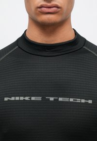 Čierny športový top s vysokým golierom, vyrobený z textúrovanej látky, s nápisom "NIKE TECH" v sivej farbe cez hruď, jemné švy.