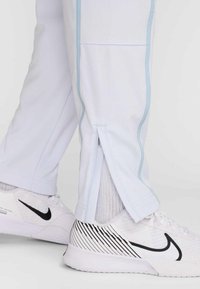 Bílé sportovní kalhoty s světle modrými akcenty a bočními rozparky na manžetách. Kombinovány s bílými teniskami, které mají strukturovanou síťovanou látku.