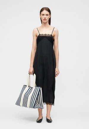 Γυναίκα που φοράει μαύρο φόρεμα με δαντέλα και κρατάει μια μεγάλη τσάντα ώμου με μαύρες και λευκές ρίγες, στέκεται μπροστά σε απλό φόντο.