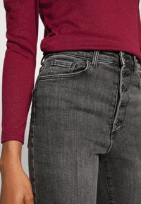 Primer plano de la cintura y cadera vistiendo unos jeans de tiro alto oscuros con cierre de botones y un suéter de manga larga burdeos, con la mano izquierda visible.