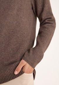Un pull en maille marron avec des poignets et un ourlet côtelés, texture douce, présentant un détail de poche à la taille, porté sur un pantalon beige clair.