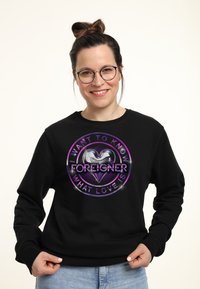 Sudadera negra con un gráfico circular que presenta un corazón plateado y el texto "QUIERO SABER QUÉ ES EL AMOR" arriba de "FOREIGNER," con acentos en púrpura y blanco.