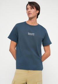 Homem a usar uma t-shirt azul com o texto branco "Leva o mundo pela cauda" e calças de carga bege, de pé contra um fundo branco liso.