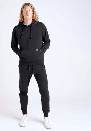 Jeune homme debout aux cheveux blonds portant un sweat à capuche noir, un pantalon de survêtement noir et des baskets blanches sur un fond clair uni.