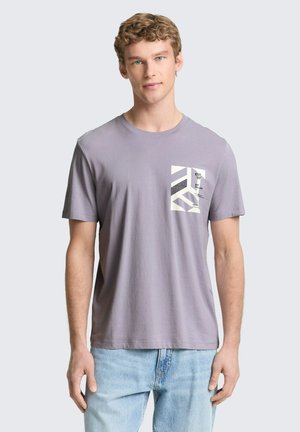 Junger Mann mit lockigem Haar, trägt ein lavendelfarbenes T-Shirt mit einer weißen geometrischen Taschenoptik und hellblaue Jeans vor einem grauen Hintergrund.