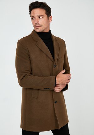 Cappotto in lana marrone con revers a contrasto, chiusura con un bottone e tasche laterali. Indossato sopra un dolcevita nero, presenta una vestibilità su misura.