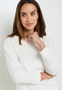 Top bianco a maniche lunghe con una texture morbida e scollo rotondo. Presenta una vestibilità rilassata e dettagli leggermente arricciati sulle spalle.