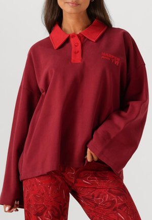 Red cotton polo-style top with a button-up collar, featuring a logo on the left side. Relaxed fit with dropped shoulders.