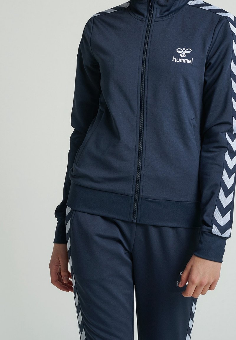 Navy urheiluharjoitusasu, jossa on valkoinen viivamalli hihoissa ja housuissa. Varustettu korkealla kauluksella ja täysipitkällä vetoketjulla. Materiaali näyttää sileältä ja joustavalta.