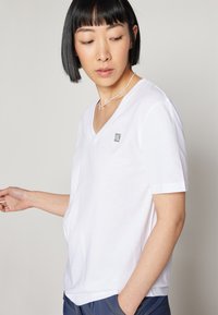 T-shirt bianco in cotone con scollo a V, maniche corte e un piccolo logo sul petto. Tessuto morbido e vestibilità rilassata.