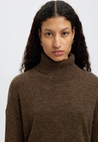 Brūns adīts augstais kakls, ar teksturētu apdari. Apkakle ir ribota, pievienojot dizainam dziļumu. Mīksta, silta materiāla izskats ir ērts.