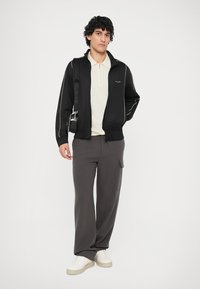 Svart hettejakke med hvite detaljer, beige polo-skjorte, grå treningsbukser og hvite sko. Modellen poserer med en crossover-veske.