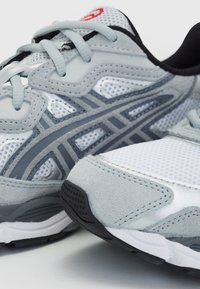 Närbild av grå och vita löparskor med svarta sulor och detaljer i nätstoff, placerade på en enkel ljus yta.