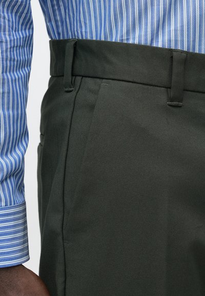 Pantalon vert foncé à texture lisse, avec des plis sur le devant et des passants pour ceinture. Associé à une chemise à manches longues bleue rayée.