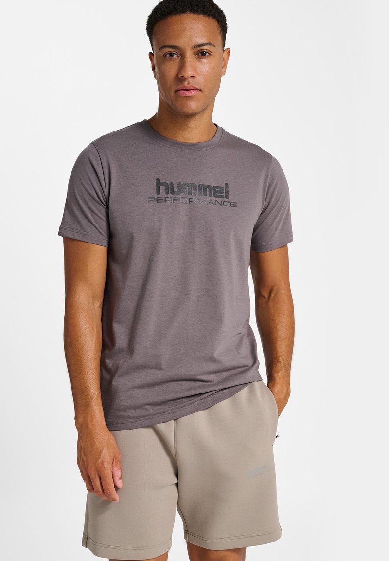 Mand iført en grå "hummel performance" t-shirt og beige shorts med den ene hånd i lommen mod en hvid baggrund.