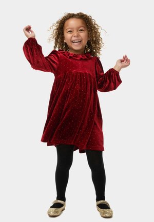 Rode fluwelen jurk met poffende mouwen en een gerimpelde halslijn, versierd met gouden stippen. Zwarte panty’s en gouden glitterballerina's maken de outfit compleet.