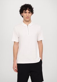 Tricou polo cu mânecă scurtă, de culoare roz deschis, cu guler cu fermoar. Material moale, croială lejeră și un design minimalist; fără modele sau grafice vizibile.