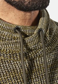 Nahaufnahme des Nackens eines Mannes, der einen strukturierten Strickpullover mit hohem Kragen und olivgrünen Kordeln trägt, die durch Metallösen gefädelt sind.