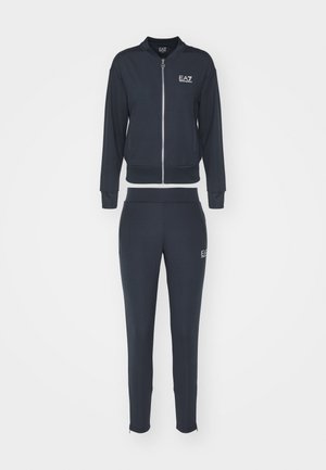 Tmavomodrý teplákový komplet pozostávajúci z bundy na zips s okrúhlym golierom a zodpovedajúcich úzkych nohavíc, pričom na oboch kúskoch sa nachádza logo.