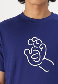 Sötétkék pamut póló, fehér absztrakt kéz grafikával az elején, amely kerekded, játékos formát mutat görbült vonalakkal.