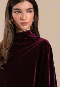 Prenda de terciopelo burdeos con un escote alto, que presenta un acento texturizado en el cuello, combinada con pendientes de forma orgánica en dorado.