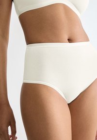 Vrouw in witte hooggeplaatste katoenen onderbroek en bijpassende beha, die het middenrif en de bovenbenen toont tegen een effen achtergrond.