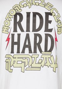 Camiseta blanca con el texto en negrita "RIDE HARD" en negro, con acentos en verde claro y relámpagos rojos, que muestra un diseño gráfico.