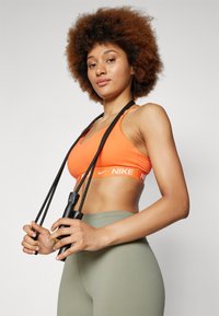 Narancssárga sportmelltartó fehér Nike logóval, olíva zöld, magas derekú leggingsszel párosítva. A felhasználó egy fekete ugrálókötéllel áll. Egyszerű háttér.