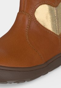 Botte en cuir marron avec une semelle foncée et un patch brillant en forme de cœur doré sur le côté supérieur près de la cheville.