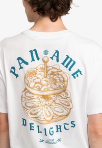 Bílá tričko s detailním zlatým graphic zobrazujícím dezertní stůl a textem "PAN AME DELIGHTS" v tyrkysové a zlaté barvě.
