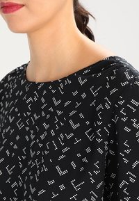 Czarna tkanina z białym geometrycznym wzorem w kropki, z szerokim dekoltem i gładką fakturą. Detal przedstawia obszar ramienia.