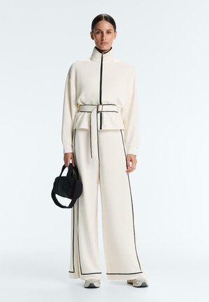 Woman wearing cream wide-leg pants and zip-up jacket with black piping and belt, holding small black handbag, standing on white background.