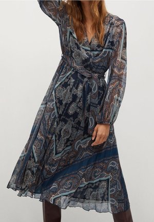 Kobieta ubrana w długą sukienkę na zakładkę z ciemnogranatowym wzorem paisley, wykonaną z przezroczystej tkaniny i z wiązaniem w talii, w połączeniu z ciemnymi kozakami sięgającymi kolan.