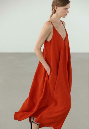 Frau geht drinnen und trägt ein fließendes ärmelloses rotes Kleid mit dünnen Trägern und schwarzen Sandalen mit hohen Absätzen.