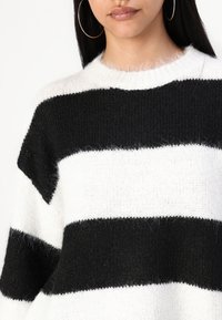 Femme portant un pull en maille duveteuse à larges rayures noires et blanches, avec de grandes boucles d'oreilles, visible du cou jusqu'au milieu du torse.