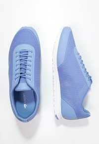 Modré sportovní boty vyrobené z prodyšné tkaniny, se zaoblenou špičkou, texturovanými detaily, plochými tkaničkami a bílou gumovou podrážkou. Logo značky je viditelné uvnitř.