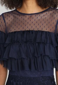 Robe bleu marine avec un haut en tulle à pois transparents et volants superposés. Le tissu est léger et texturé, agrémenté d'accents en tissu à la taille.