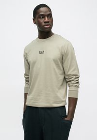 Μπεζ μπλούζα με μακρύ μανίκι και στρογγυλή λαιμόκοψη, με μικρό μαύρο λογότυπο EA7 στο στήθος. Το υλικό φαίνεται μαλακό με λείο υφή.