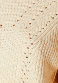 Kermankollainen neulepaita, jossa on teksturoitu kuvio pienillä ruskeilla tähdenmuotoilla, hienovaraiset aukot yksityiskohdat ja ribbireunat.